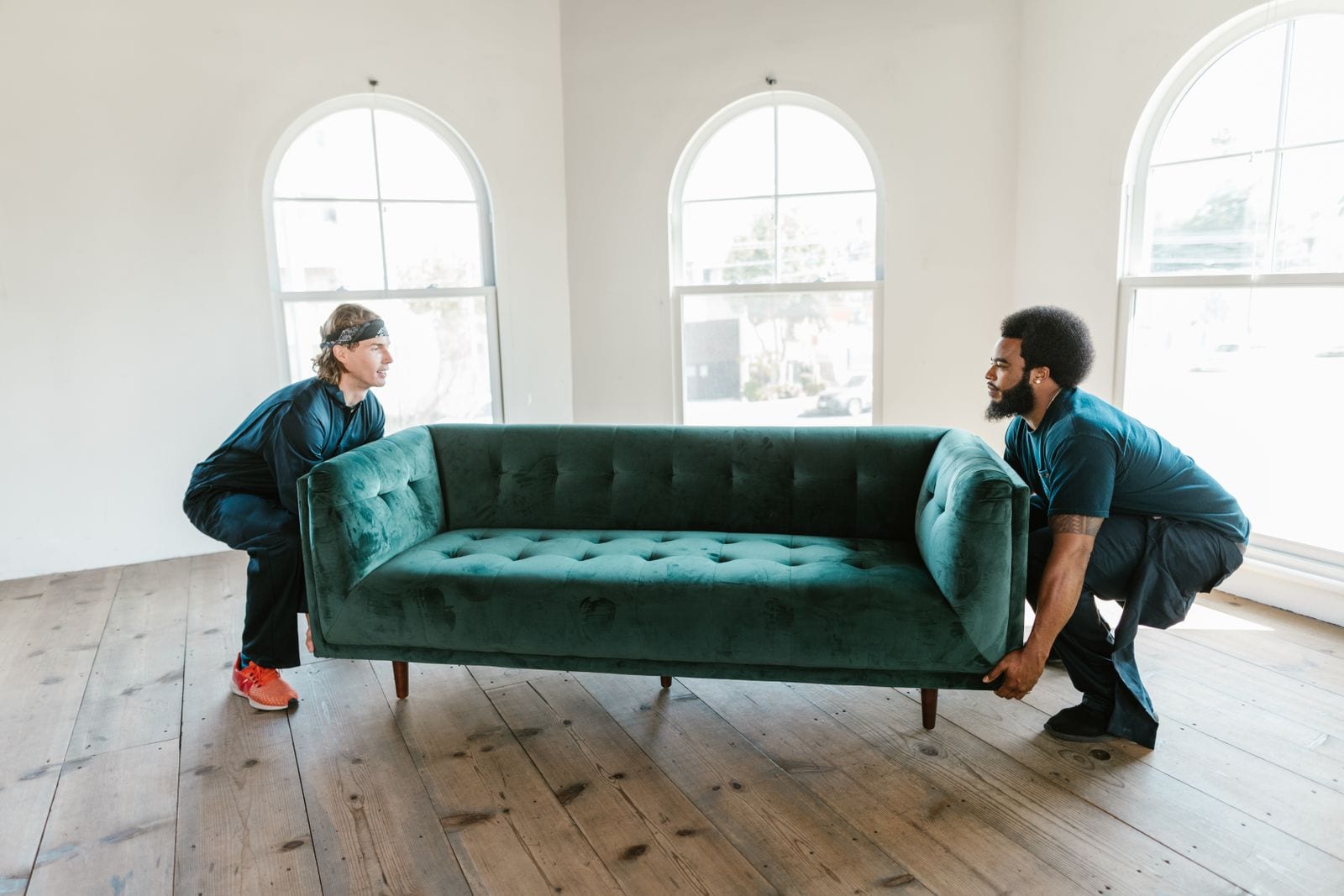 Two movers carefully lifting a green velvet sofa together in a sunlit, hardwood-floored room.